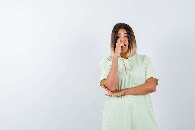 Niña mordiendo los dedos emocionalmente, sosteniendo la mano debajo del codo en camiseta y mirando asustada. vista frontal.