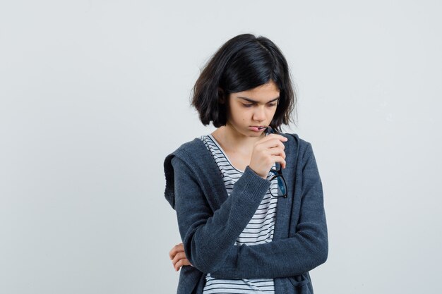 Niña mordiendo vasos en camiseta, chaqueta y mirando deprimido.