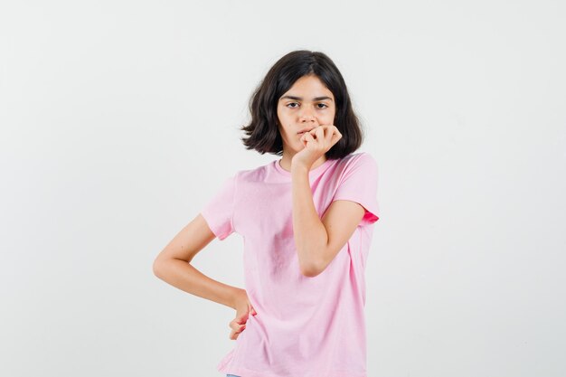 Niña mordiéndose las uñas mientras piensa en camiseta rosa y parece preocupado, vista frontal.