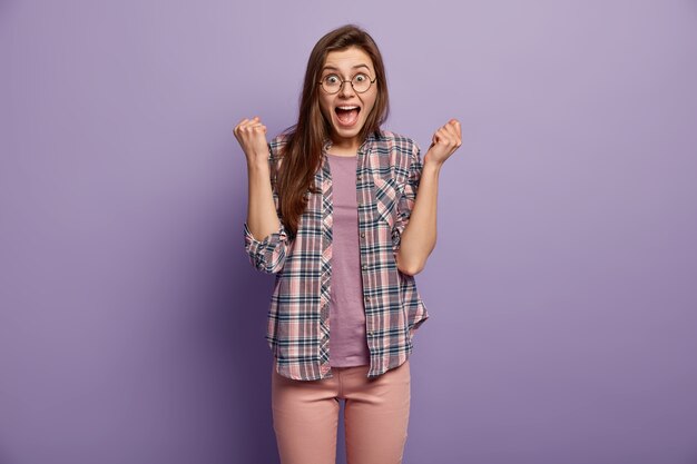 La niña morena feliz aprieta los puños como la ganadora, ha sorprendido la expresión facial llena de alegría, mantiene la boca abierta, usa una elegante camisa a cuadros, posa y gestos sobre la pared púrpura, obtuvo la victoria