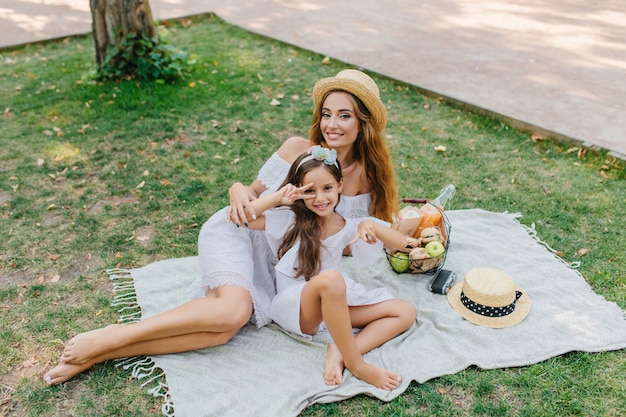 Niña morena posando sobre una manta mostrando el signo de la paz con una sonrisa emocionada. Retrato al aire libre de una mujer bonita y su hija tirada en el suelo con una cesta de manzanas.