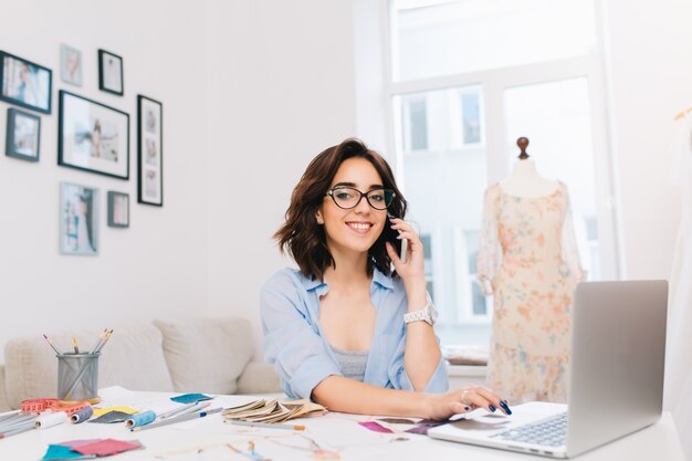 Una niña morena sonriente con una camisa azul está sentada a la mesa en el estudio. Ella está hablando por teléfono y escribiendo en la computadora. Ella sonríe a la cámara.