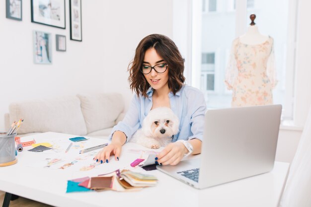 Una niña morena sonriente con una camisa azul está sentada a la mesa en el estudio. Ella trabaja con cosas creativas. Tiene un lindo perro de rodillas.