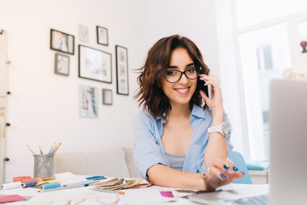 Una niña morena sonriente con una camisa azul está trabajando en el taller. Ella está hablando por teléfono y escribiendo en la computadora. Ella sonríe a la cámara.