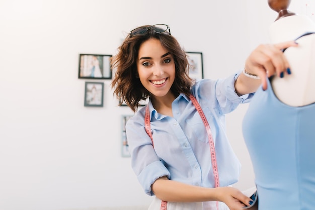 Una niña morena sonriente está creando un vestido azul en el maniquí. Ella trabaja con una camisa azul. Es un retrato de una niña en el estudio del taller.