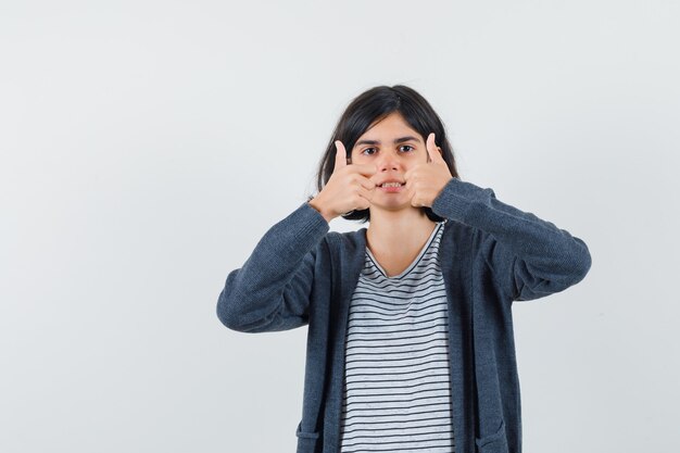 Niña mostrando doble pulgar hacia arriba en camiseta, chaqueta y mirando confiado,