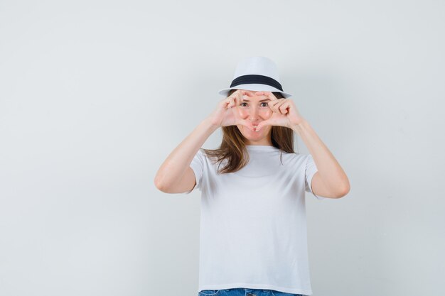 Niña mostrando gesto de corazón en camiseta blanca, sombrero y mirando alegre. vista frontal.