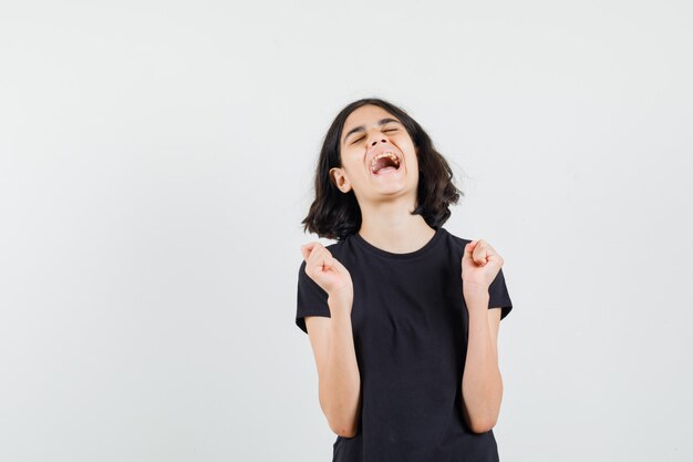 Niña mostrando gesto de éxito en camiseta negra y mirando feliz. vista frontal.