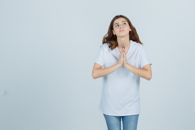 Niña mostrando gesto de namaste en camiseta blanca y mirando esperanzado, vista frontal.