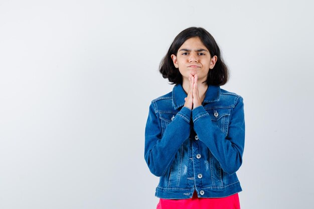 Niña mostrando gesto de namaste en camiseta roja y chaqueta de jean y mirando seria, vista frontal.