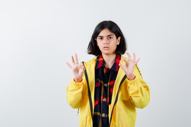 Niña mostrando gesto ok en camisa a cuadros, chaqueta y mirando confiado