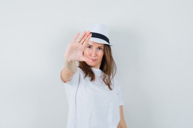 Niña mostrando gesto de parada con sombrero de camiseta blanca y mirando confiado