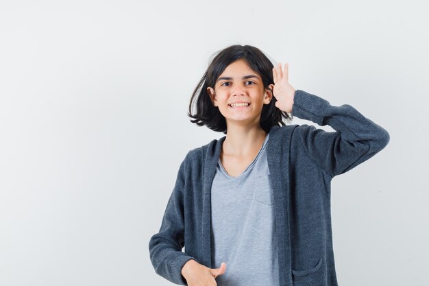 Niña mostrando gesto de saludo en camiseta, chaqueta y mirando alegre, vista frontal.