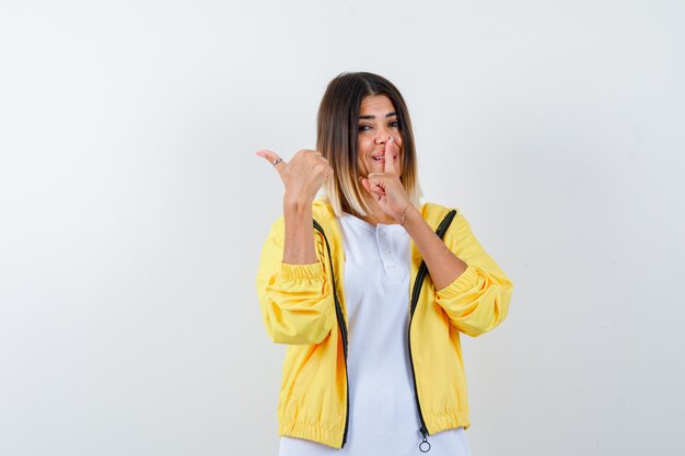 Niña mostrando gesto de silencio, apuntando a un lado con el pulgar en camiseta blanca, chaqueta amarilla y mirando alegre, vista frontal.