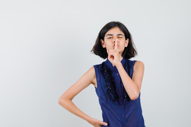 Niña mostrando gesto de silencio en blusa azul y mirando con cuidado.