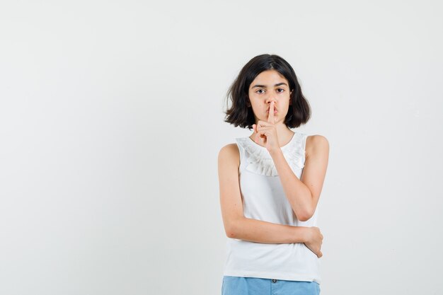 Niña mostrando gesto de silencio en blusa blanca, pantalones cortos y mirando con cuidado. vista frontal.