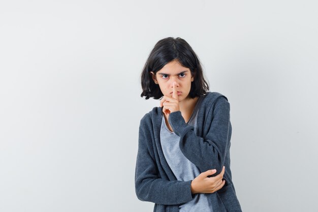 Niña mostrando gesto de silencio en camiseta, chaqueta y mirando serio