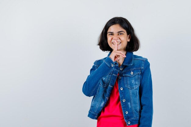 Niña mostrando gesto de silencio en camiseta roja y chaqueta de jean y mirando feliz