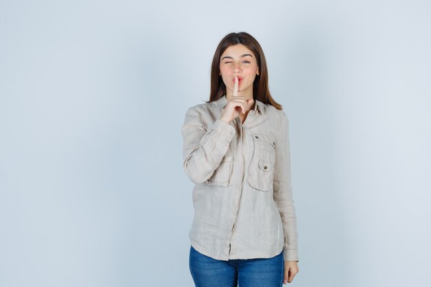 Niña mostrando gesto de silencio, guiñando un ojo en camisa beige, jeans y luciendo linda. vista frontal.