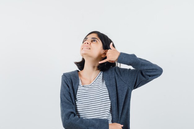 Niña mostrando gesto de teléfono en camiseta, chaqueta y aspecto soñador.