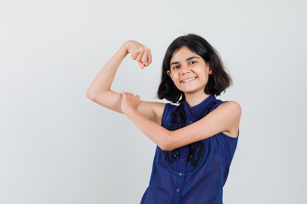 Niña mostrando los músculos del brazo en blusa azul y mirando confiada.