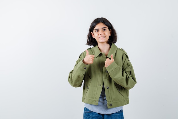 Niña mostrando los pulgares para arriba con ambas manos en suéter gris, chaqueta de color caqui, pantalón de mezclilla y mirando feliz, vista frontal.