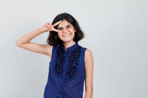 Niña mostrando el signo v cerca del ojo en blusa azul y luciendo juguetón.