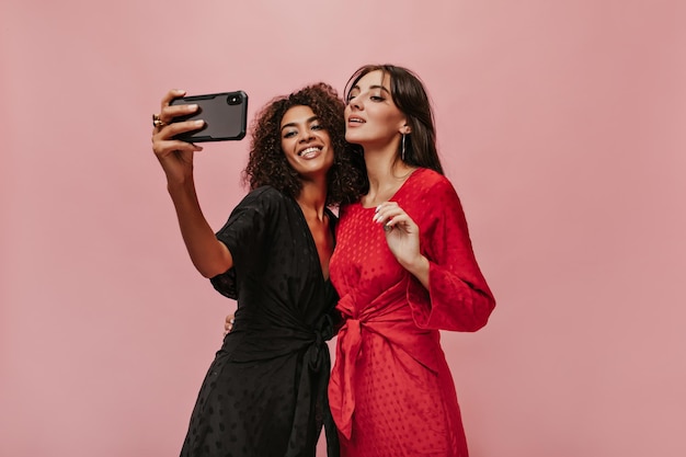 Niña mulata sonriente con peinado rizado en vestido negro tomando fotos con su amiga moderna con cabello largo en vestido brillante