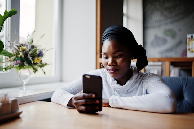 Niña musulmana africana con hiyab negro sentada en un café con teléfono móvil a mano