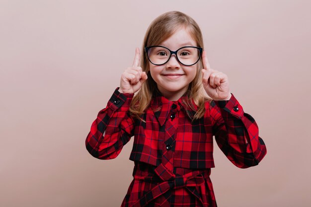 Niña muy inteligente de pie sobre una pared beige sosteniendo el dedo grande con una sonrisa