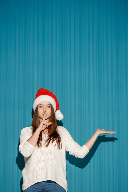La niña de Navidad sorprendida con un gorro de Papá Noel que muestra a la derecha sobre el fondo azul.