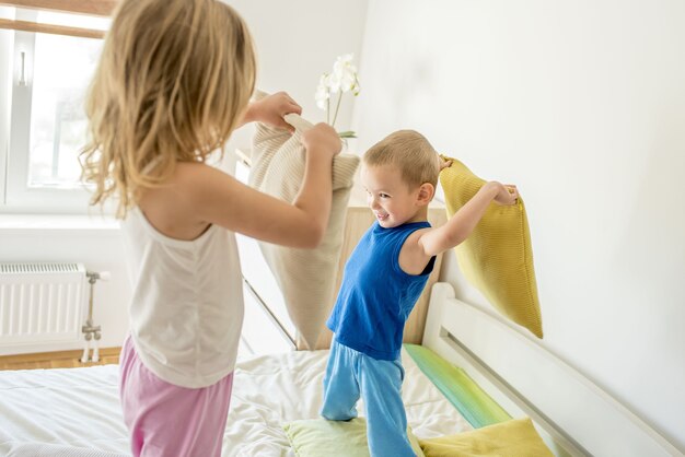 Niña y un niño sonriendo y teniendo una pelea de almohadas en una cama