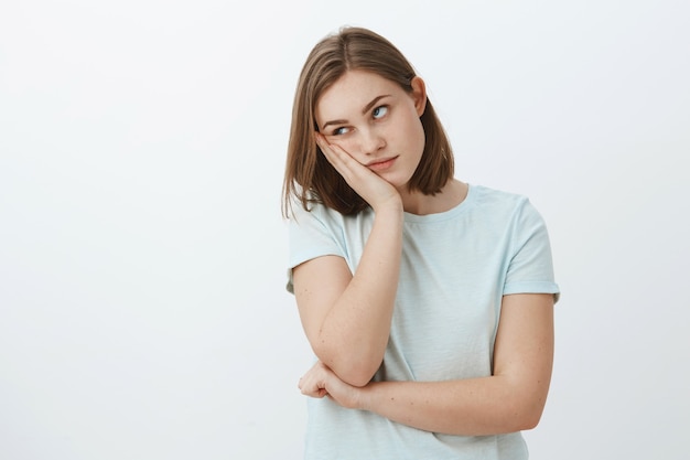 La niña odia asistir a clases aburridas en la universidad, con la cara inclinada en la mano, poniendo los ojos en blanco por el aburrimiento y la molestia, de pie indiferente y disgustado sobre la pared gris con una camiseta de moda