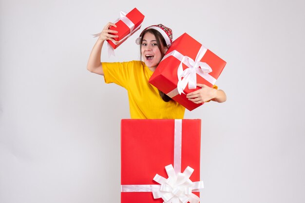 Niña de ojos abiertos con gorro de Papá Noel sosteniendo regalos de pie detrás de un gran regalo de Navidad en blanco