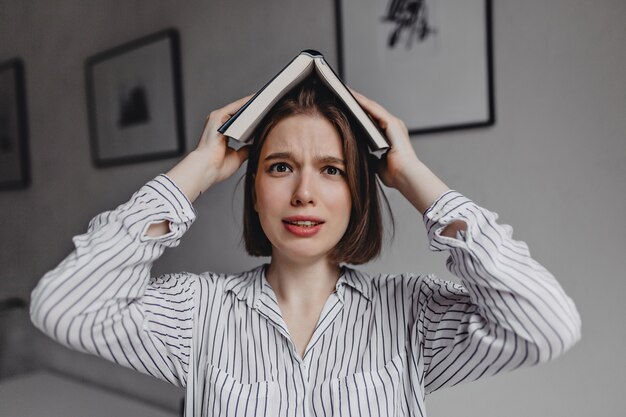 La niña de ojos marrones se pone el libro en la cabeza, mirando a la cámara desconcertada y asustada.