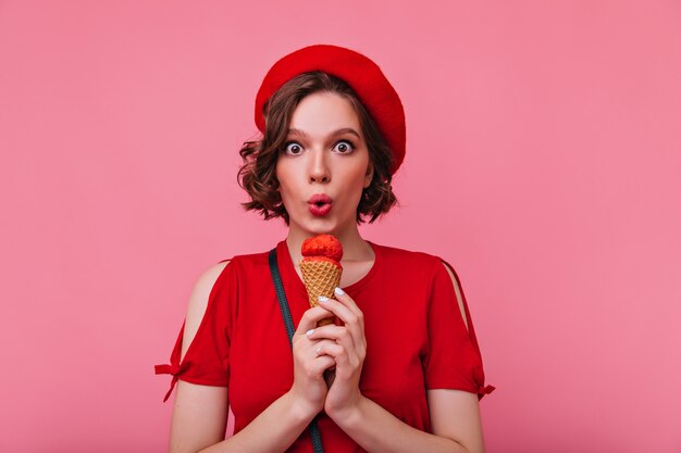 Niña de ojos marrones soñadora comiendo helado con expresión de cara de sorpresa. Impresionante mujer con corte de pelo corto viste boina roja.