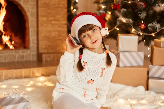 Niña optimista con suéter blanco y sombrero de santa claus, mirando a la cámara, teniendo un ambiente festivo, tomando por teléfono, sentada en el piso cerca del árbol de Navidad, cajas de regalo y chimenea.