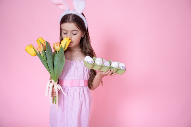 Niña con orejas de conejo de Pascua y una bandeja de huevos en sus manos oliendo un ramo de tulipanes sobre un fondo de estudio rosa