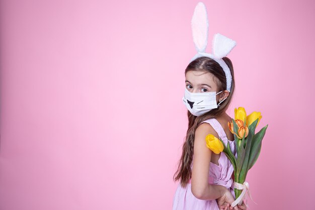 Niña con orejas de conejo de pascua y vistiendo una mascarilla médica sostiene un ramo de tulipanes en sus manos en un rosa