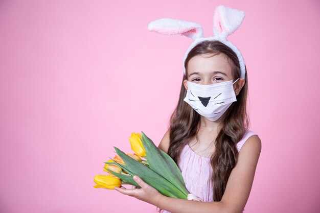 Niña con orejas de conejo de pascua y vistiendo una mascarilla médica sostiene un ramo de tulipanes en sus manos sobre un fondo de estudio rosa