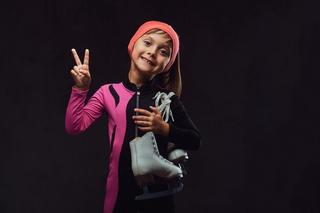 Una niña patinadora feliz vestida con ropa deportiva sostiene patines de hielo en un hombro y muestra la mano de la victoria. Aislado en un fondo de textura oscura.