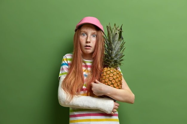 La niña pecosa asustada emocional abraza la piña, le gustan las frutas tropicales, usa gorra y camiseta a rayas, tiene un brazo roto, aislado en una pared verde. Concepto de infancia y estilo de vida.