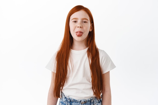 Niña pelirroja con pelo largo rojo natural y pecas, muestra lengua tonta e infantil, divirtiéndose. Niño feliz con peinado de jengibre sintiéndose positivo, pared blanca