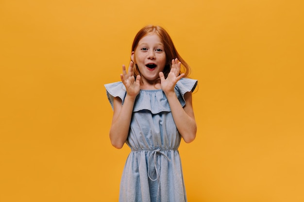 Niña pelirroja sorprendida con vestido azul se regocija con el fondo naranja Retrato de niña feliz con traje de mezclilla mira a la cámara