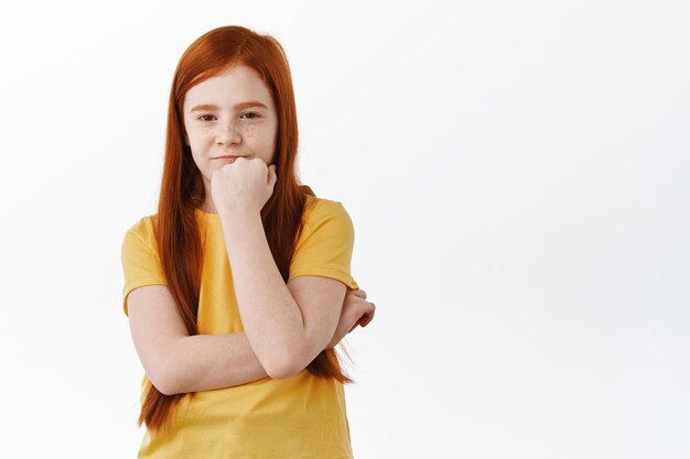 Niña pelirroja sospechosa que tiene dudas, mirando con incredulidad y cara vacilante a la cámara, pensando, de pie sobre fondo blanco