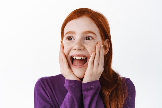 La niña pelirroja se ve sorprendida y feliz a un lado, asombrada con algo súper genial, sonriendo impresionada o fascinada, mira con admiración la pancarta, la pared blanca