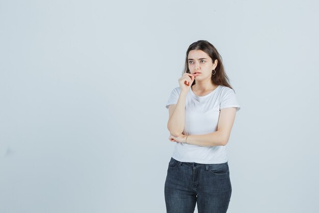 Niña pellizcando el labio inferior en camiseta, jeans y mirando pensativo. vista frontal.