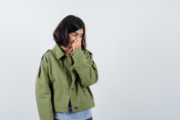 Niña pellizcando la nariz debido al mal olor en suéter gris, chaqueta caqui, pantalón de mezclilla y mirando acosado, vista frontal.