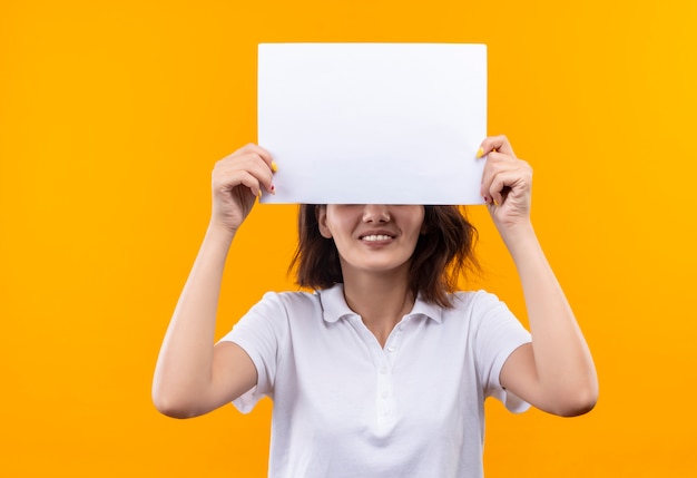 Foto gratuita niña con pelo corto vistiendo polo blanco escondiendo ojos beahind página en blanco sonriendo alegremente