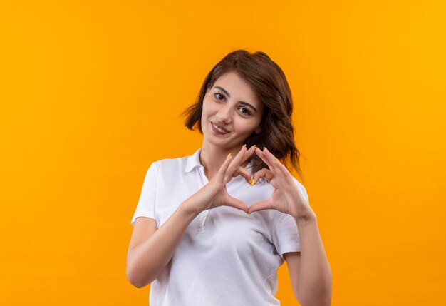 Niña con pelo corto vistiendo polo blanco haciendo gesto de corazón con los dedos sobre el pecho siendo encantador sonriendo